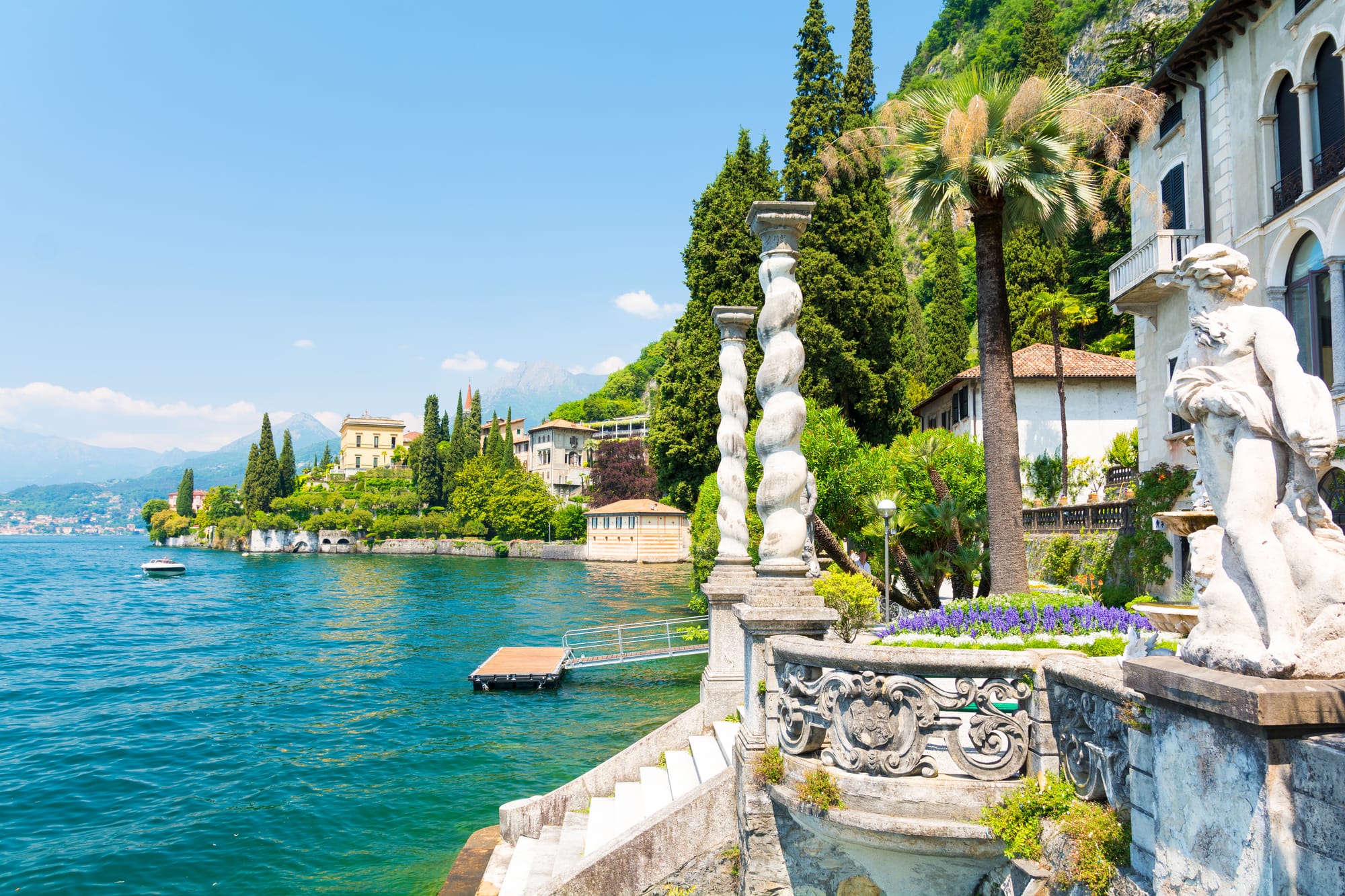 The magnificent botanical gardens of a Lake Como villa, with colorful flowers and manicured lawns leading to a stunning view of the lake and surrounding mountains. A key attraction for any Lake Como travel guide.