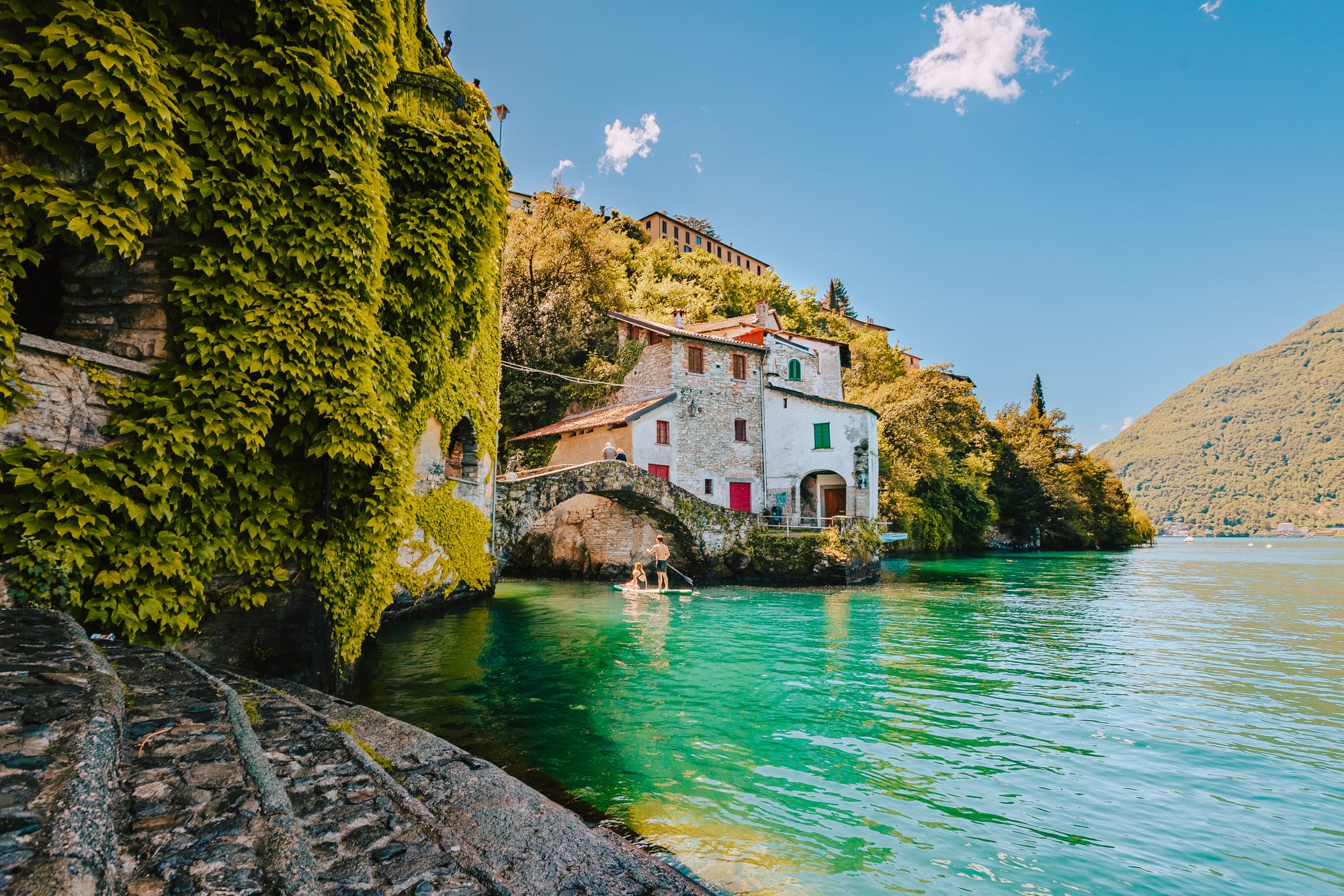 The charming village of Nesso on Lake Como, Italy, featuring the ancient stone Civera Bridge arching over the dramatic Orrido di Nesso gorge and waterfall.