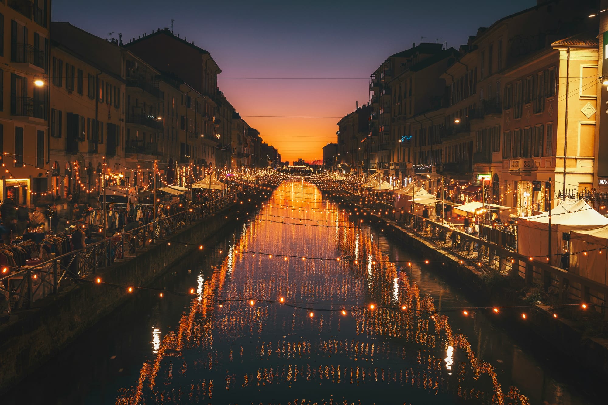 The lively Naviglio Grande canal at night in Milan, with warm lights from bustling aperitivo bars and restaurants beautifully reflecting on the water's surface.