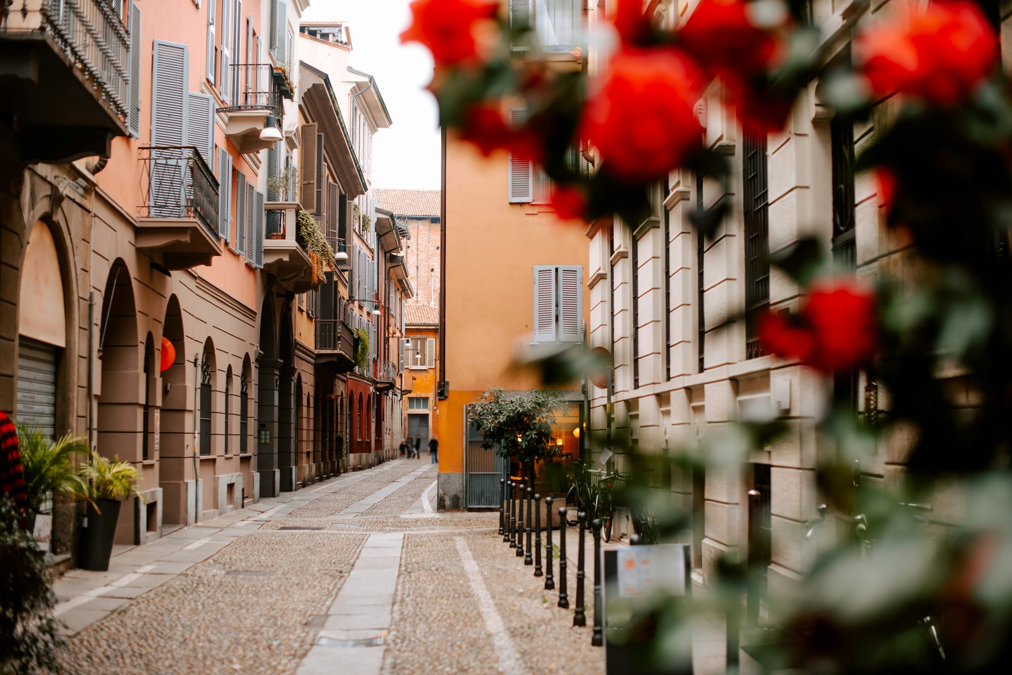 A charming cobblestone street in Milan's historic Brera district, lined with ivy-clad buildings, art galleries, and outdoor café tables, showcasing the neighborhood's bohemian atmosphere.