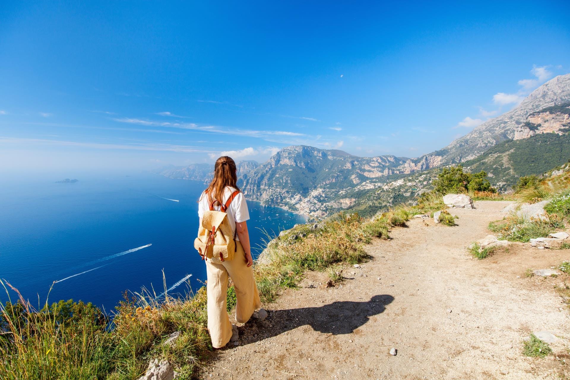 A young woman hiking the scenic Path of the Gods trail with a panoramic view of the mountains and the sea.