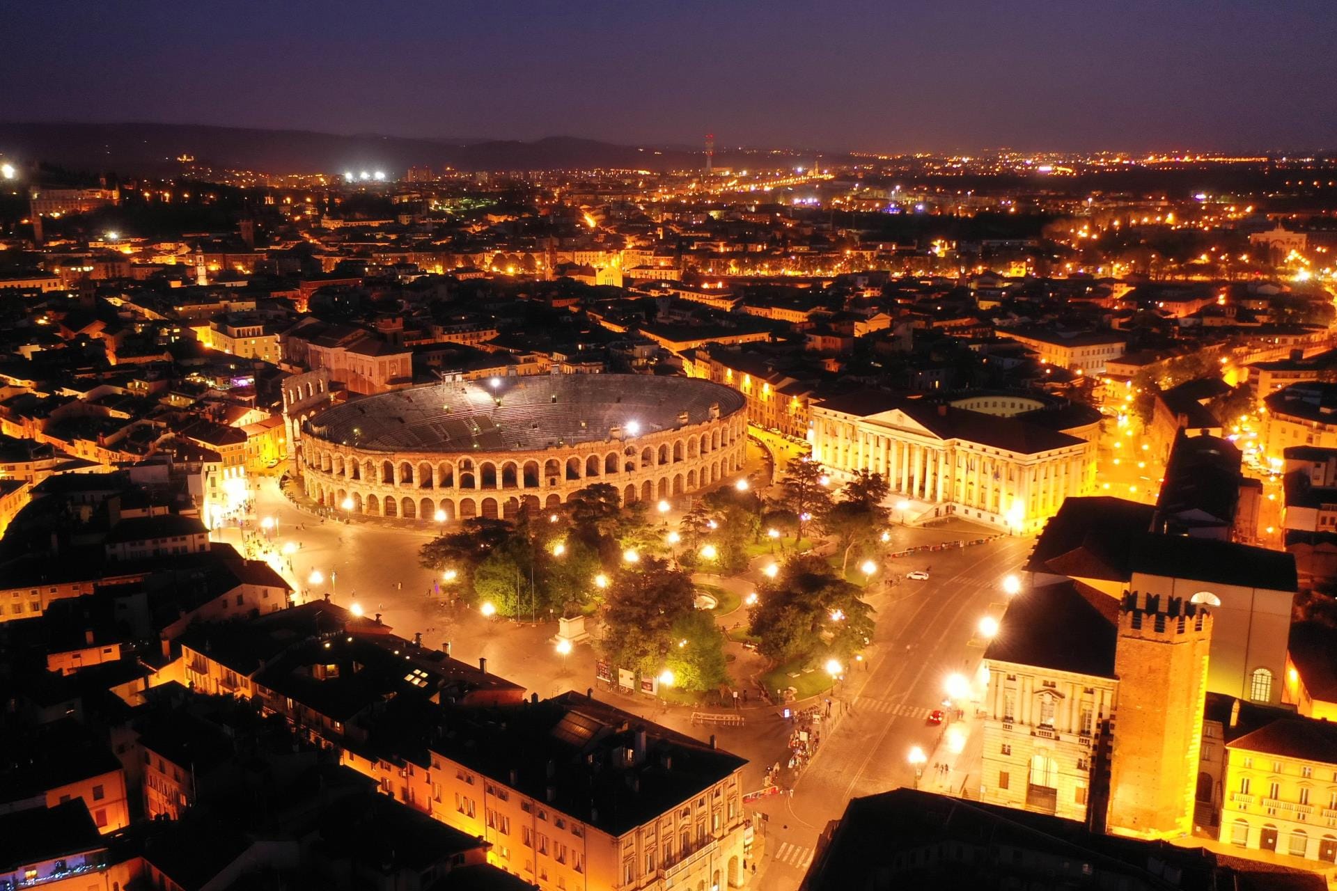An aerial night drone shot of the illuminated Verona Arena and City Hall in Piazza Bra, the designated venue for the Milano Cortina 2026 closing ceremony.