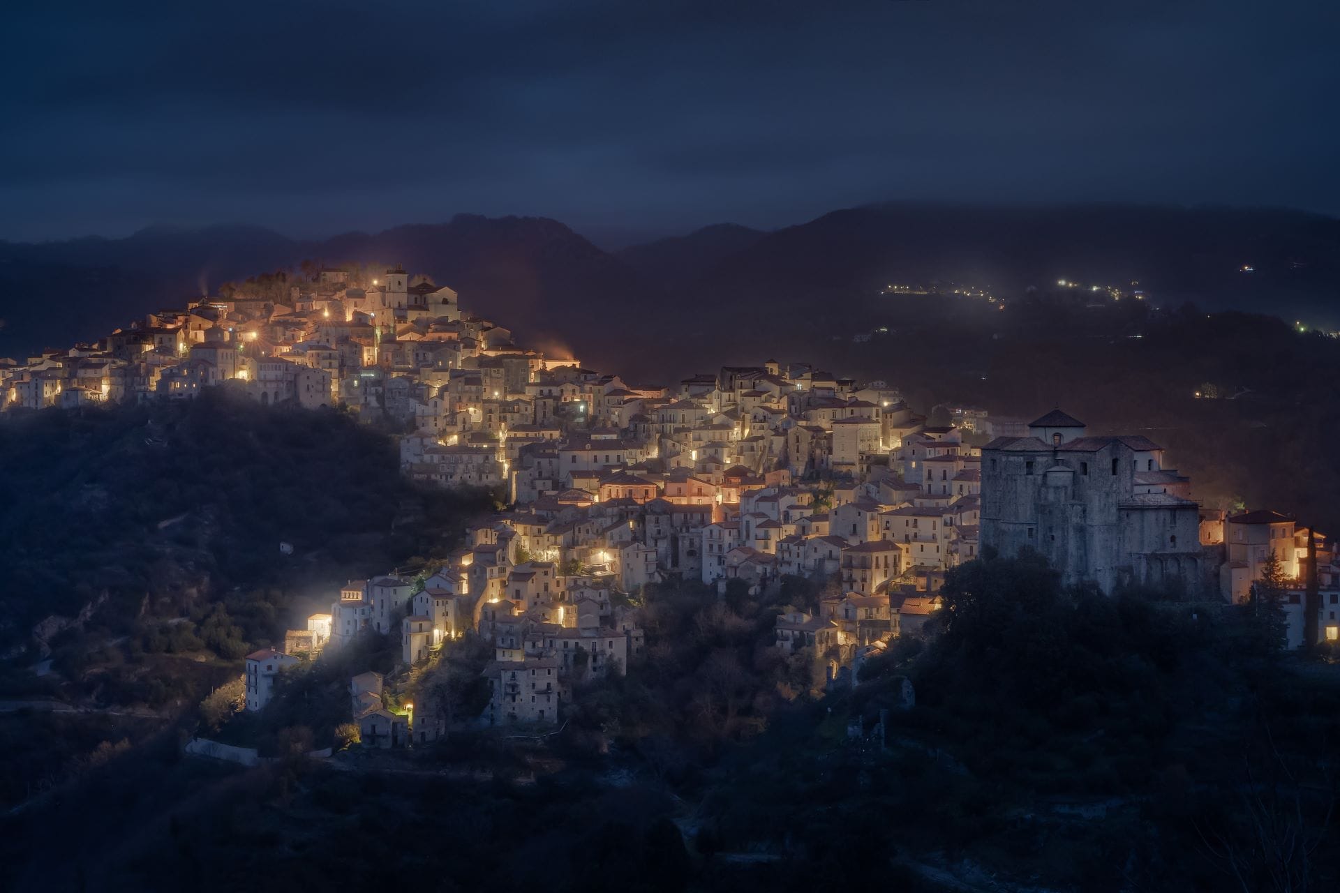 A long-exposure evening photograph of the hilltop village of Rivello in Basilicata, showing the illuminated streets cascading down three hills against the dark silhouette of the Sirino Massif.
