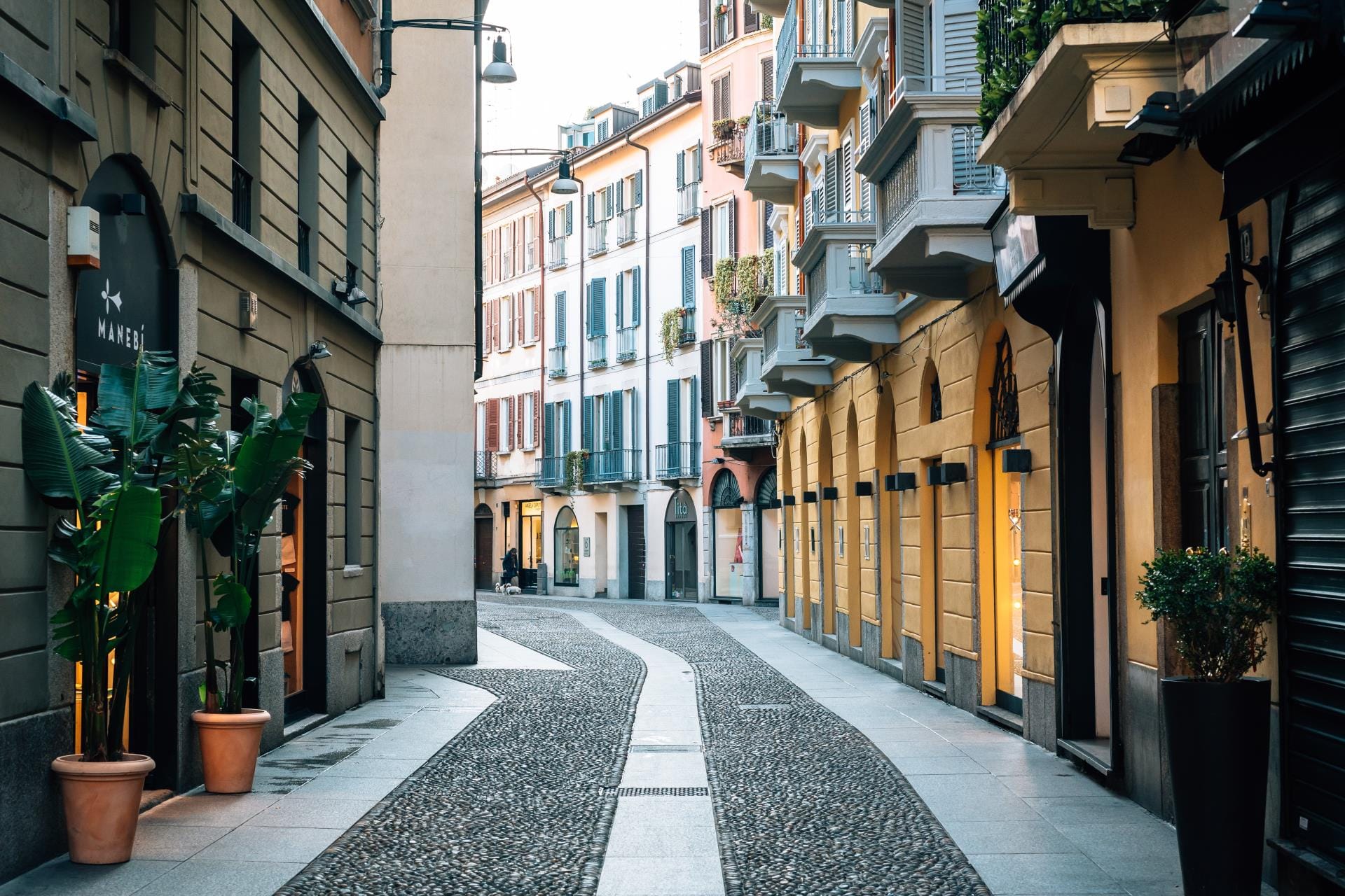 A charming and colorful narrow cobblestone street in the historic Brera district of Milan, Italy, featuring ivy-clad buildings and boutique storefronts.