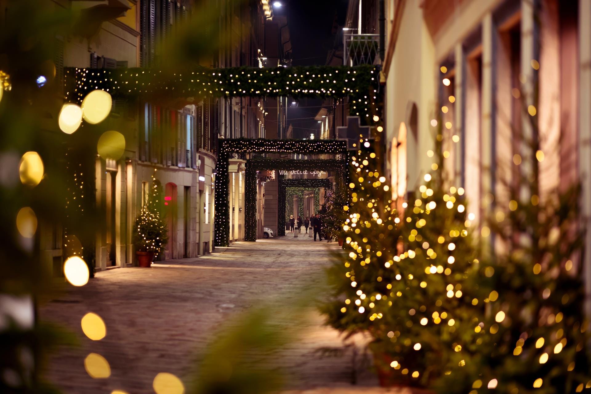 Night view of Via della Spiga in Milan decorated with festive Christmas lights and holiday displays in luxury fashion boutique windows.