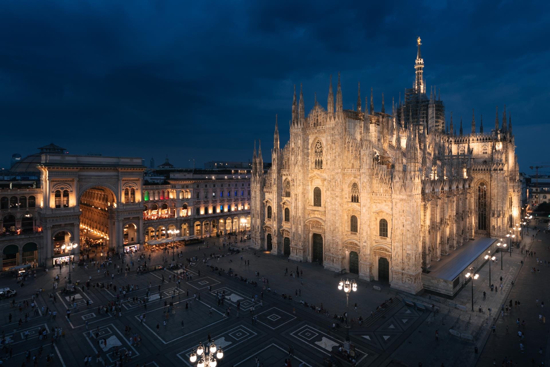 A wide night-time shot of Piazza del Duomo, showing the illuminated facade of the Milan Cathedral and the glowing entrance of the Galleria Vittorio Emanuele II.