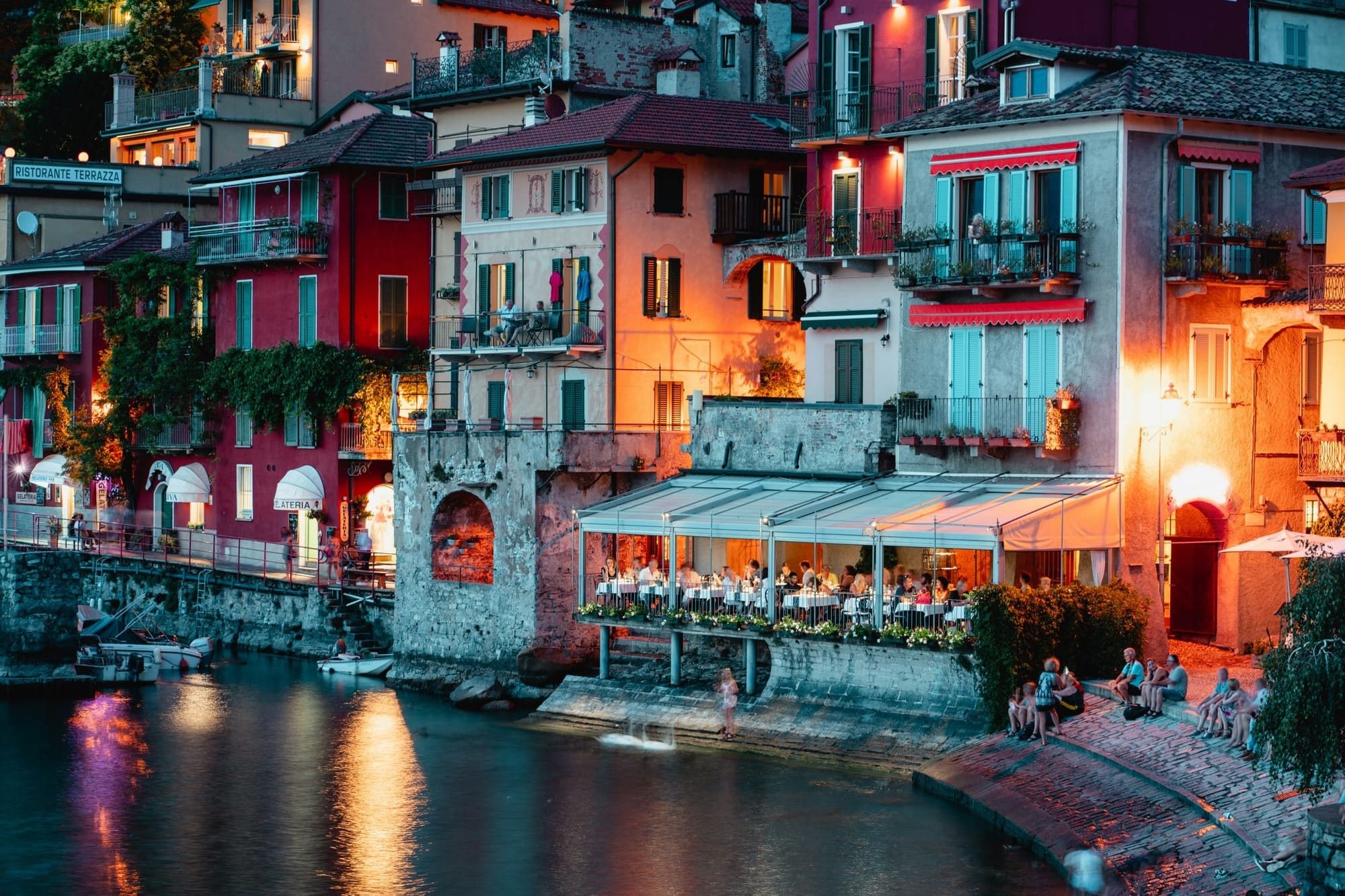 Romantic evening view of Varenna waterfront with warm lights from lakeside restaurants reflecting on the surface of Lake Como at dusk.