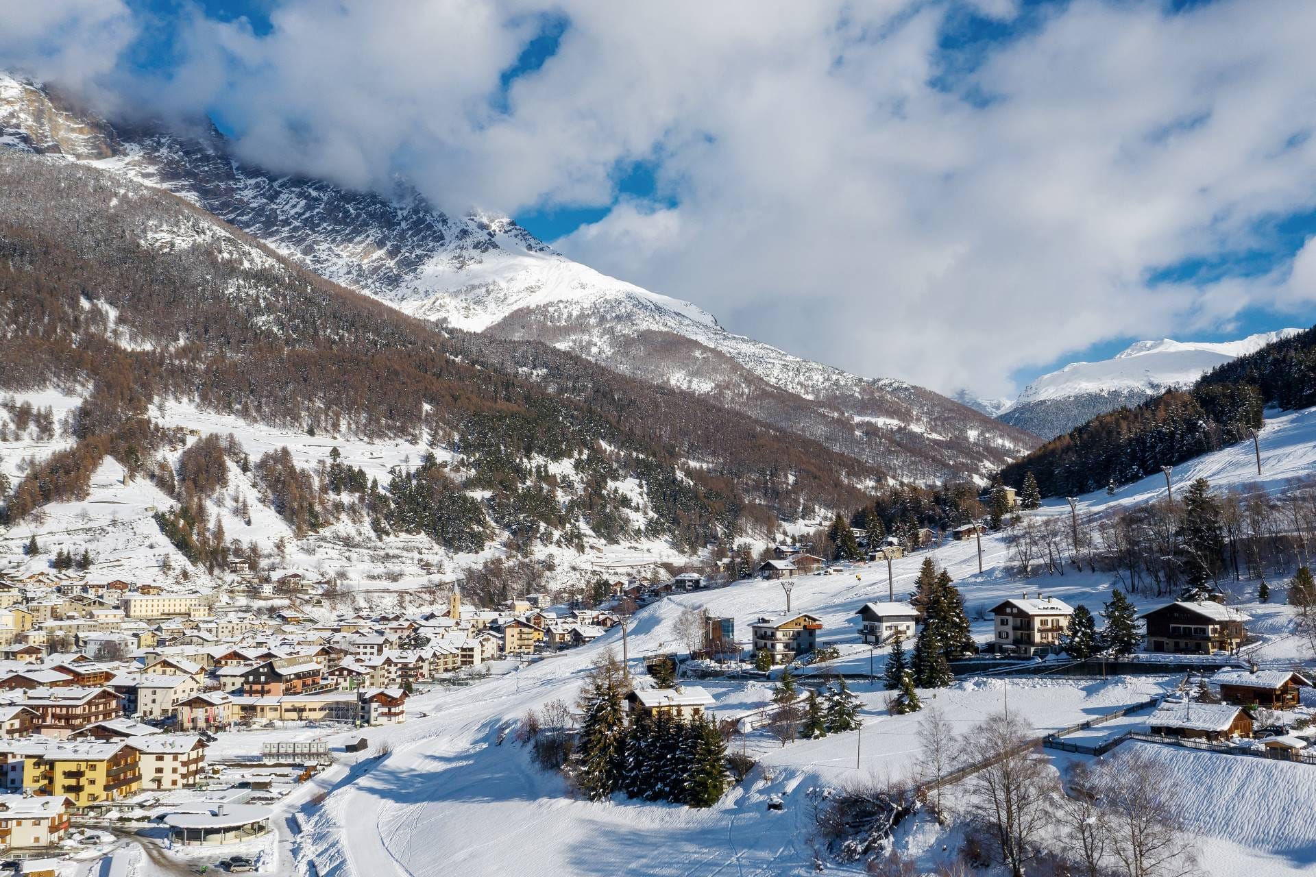 Aerial drone shot of the medieval town center of Bormio covered in snow, surrounded by the Rhaetian Alps at twilight.