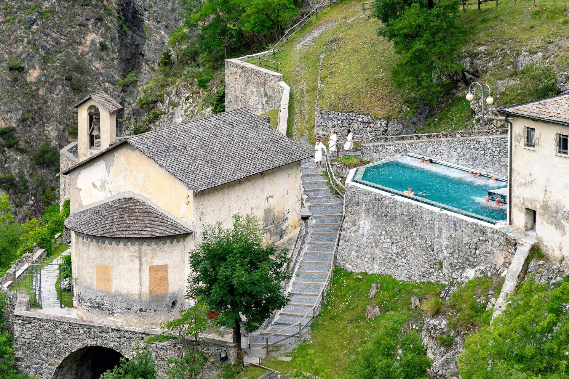 Aerial view of the outdoor panoramic thermal pool at QC Terme Bagni Vecchi in Bormio with steam rising against the snowy Alps.