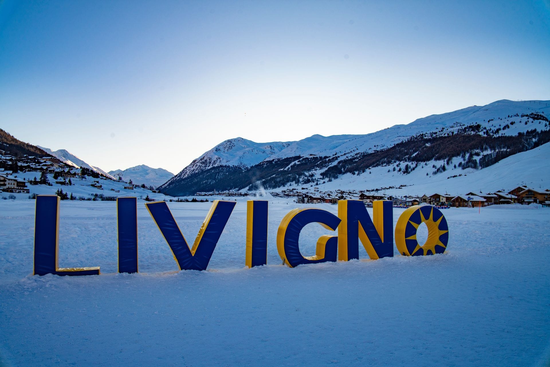 Large wooden Livigno sign covered in snow with alpine mountains in the background.