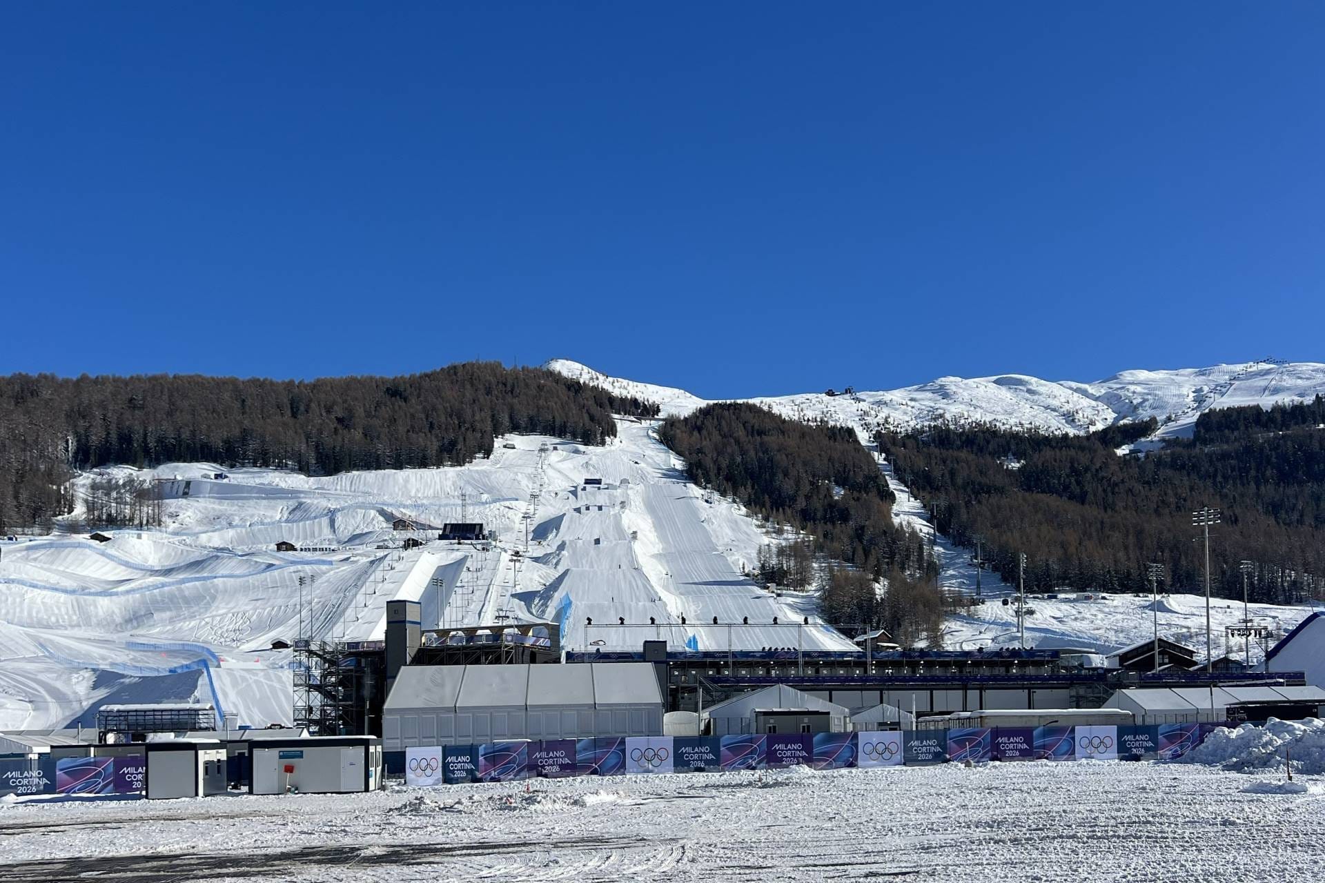 Panoramic view of the snow sports competition venues and olympic infrastructure in Livigno, Italy, featuring groomed slopes, spectator stands, and security checkpoints prepared for the 2026 winter games.
