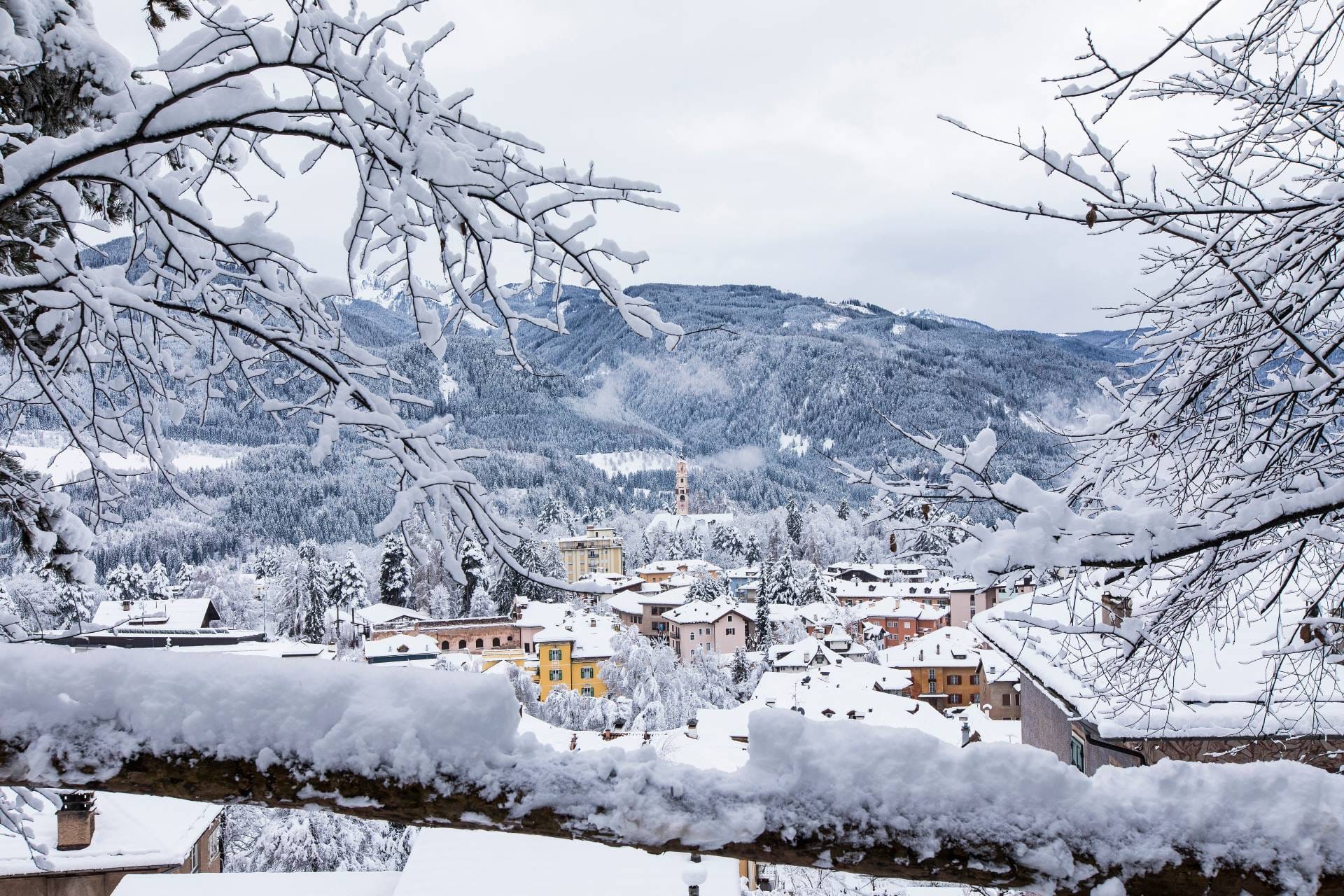 A panoramic winter view of the town of Cavalese in Val di Fiemme, showing traditional alpine architecture under a blanket of snow.