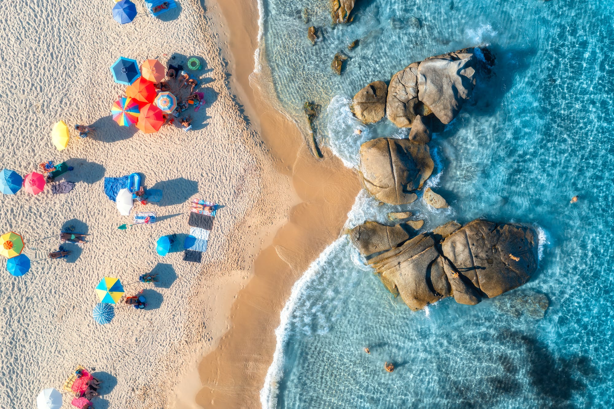 Aerial drone view of a beautiful sandy beach in Italy with colorful umbrellas, turquoise water, and rocks, representing a perfect Italian coast vacation.