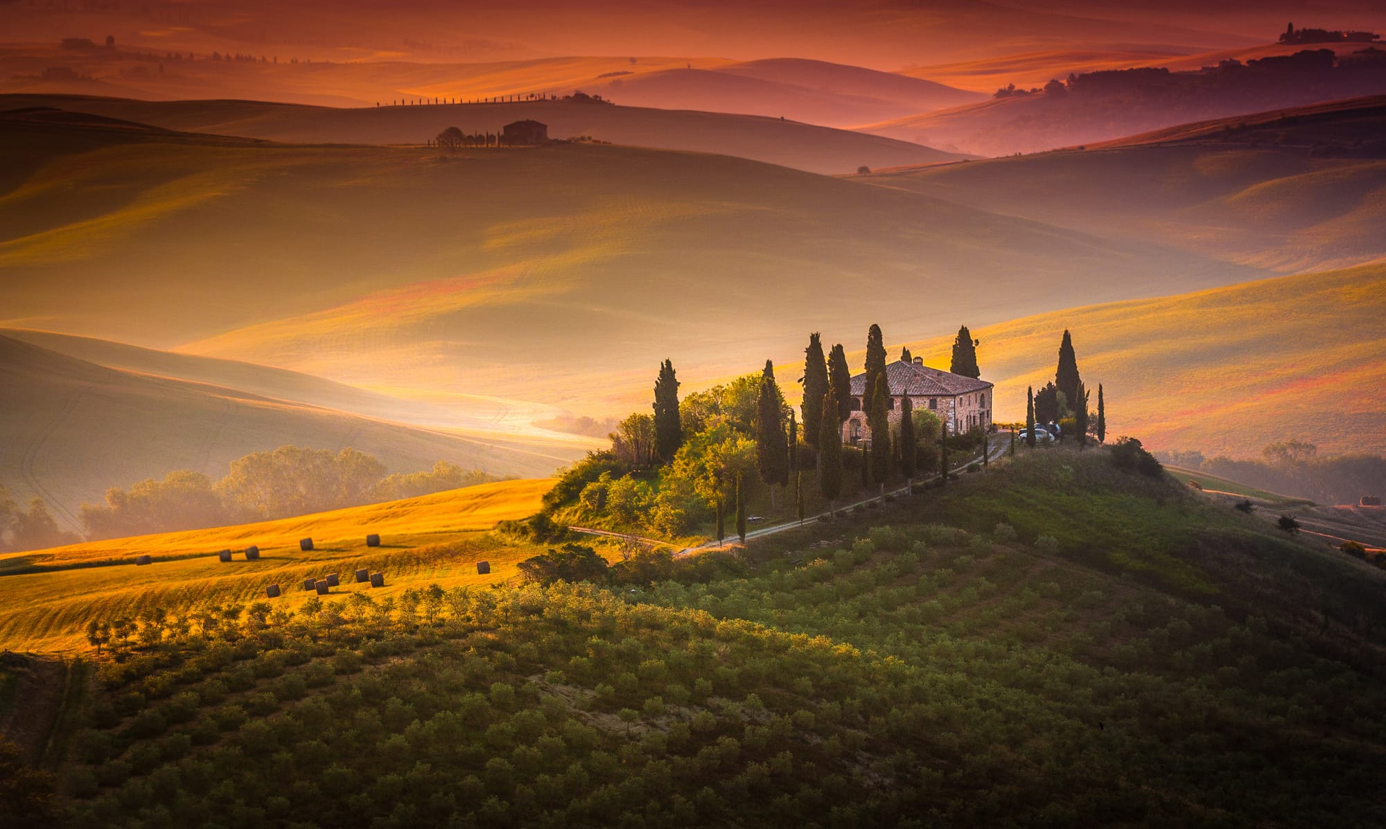 Aerial view of a stone Tuscan farmhouse surrounded by rolling hills and cypress trees in Val d'Orcia at sunset.