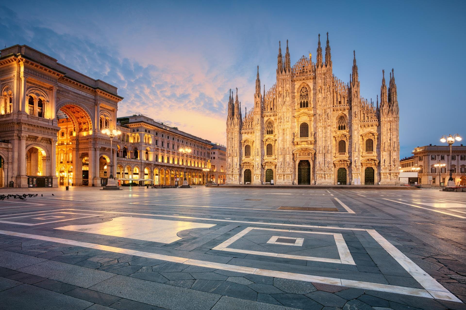 A breathtaking sunrise view of the Duomo di Milano cathedral with the entrance of the Galleria Vittorio Emanuele II visible on the left under a soft morning sky.